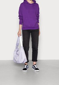 Felpa viola con tasca frontale, jeans neri e sneakers neri con dettagli blu. Borsa grigia con una tracolla ampia.