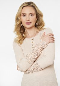Hosszú ujjú, bordázott felső világos bézs színben, csipkés díszítéssel az ujjak végén és díszgombokkal az elején. Textúrált anyag és fodros nyakvonal.