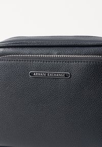 Schwarze, strukturierte Lederhandtasche mit Reißverschluss, ausgestattet mit einer schwarzen Metall-Logo-Plakette, die "ARMANI EXCHANGE" liest. Einfaches und minimalistisches Design.