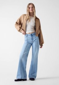 Jean flare bleu clair avec coutures avant, associé à une veste courte beige et un top blanc. Le denim présente une texture légèrement usée.