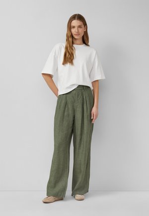 Woman with long light brown hair wearing a white oversized t-shirt, loose olive green pants, and beige shoes standing against a gray wall.