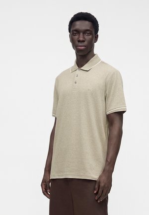 Jonge man die een beige korte mouwen polo met knopen draagt, staand tegen een effen witte achtergrond.