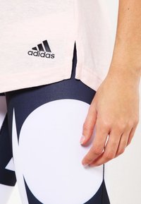 Detailní záběr ruky poblíž legín s námořnickým a bílým vzorem a světle růžového trička Adidas s postranním rozparkem.