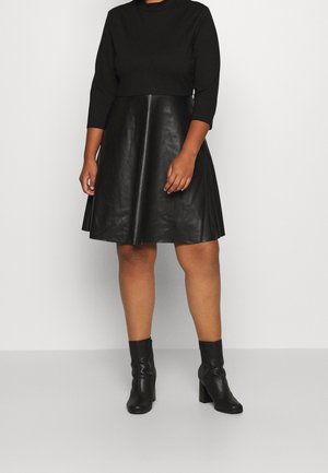 Persona con una camiseta negra de manga larga, falda negra de cuero sintético y botas negras con tacón hasta la mitad de la pantorrilla, de pie frente a un fondo liso.
