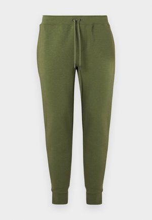 Homme portant une casquette marron, un t-shirt blanc à manches courtes avec un logo, un pantalon de jogging vert olive et des baskets blanches, debout les mains dans les poches.