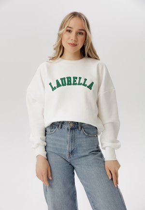Biały sweter z zielonym haftem z napisem "LAURELLA" łukiem na piersi, w połączeniu z jeansami o wysokim stanie w kolorze niebieskim. Miękka tkanina, luźny krój.