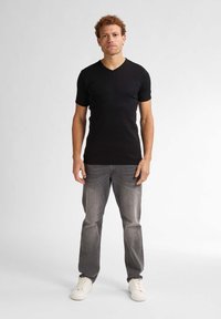 Un tricou negru cu decolteu în V, cu mâneci scurte, purtat cu blugi gri și adidași albi. Modelul stă în picioare pe un fundal neutru.