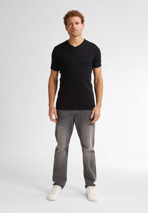 Camiseta negra de cuello en V, de mangas cortas, combinada con jeans grises y zapatillas blancas. El modelo está de pie frente a un fondo neutro.