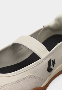 Chaussure blanche à enfiler avec une bride élastique, une étoile noire et un logo chevron, une semelle intérieure noire et un bord de semelle marron visible.