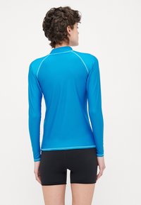 T-shirt atlético de mangas longas em tecido azul brilhante com uma textura suave, apresentando costuras em contraste. Combinado com calções pretos.