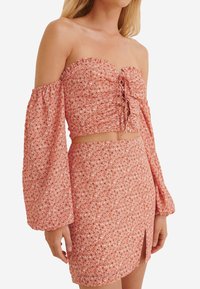 Rose-patterned, off-the-shoulder crop top with ruffled edges, long bell sleeves, and a lace-up front. Paired with a matching fitted mini skirt.