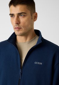 Veste zippée marine en tissu doux avec un col droit. Présente un petit logo "GUESS" brodé sur le côté gauche.