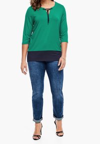 Maglietta verde a maniche lunghe con un pannello inferiore blu navy, caratterizzata da scollo a V e dettaglio con fiocco. Abbinata a jeans in denim blu con risvolto e tacchi neri.