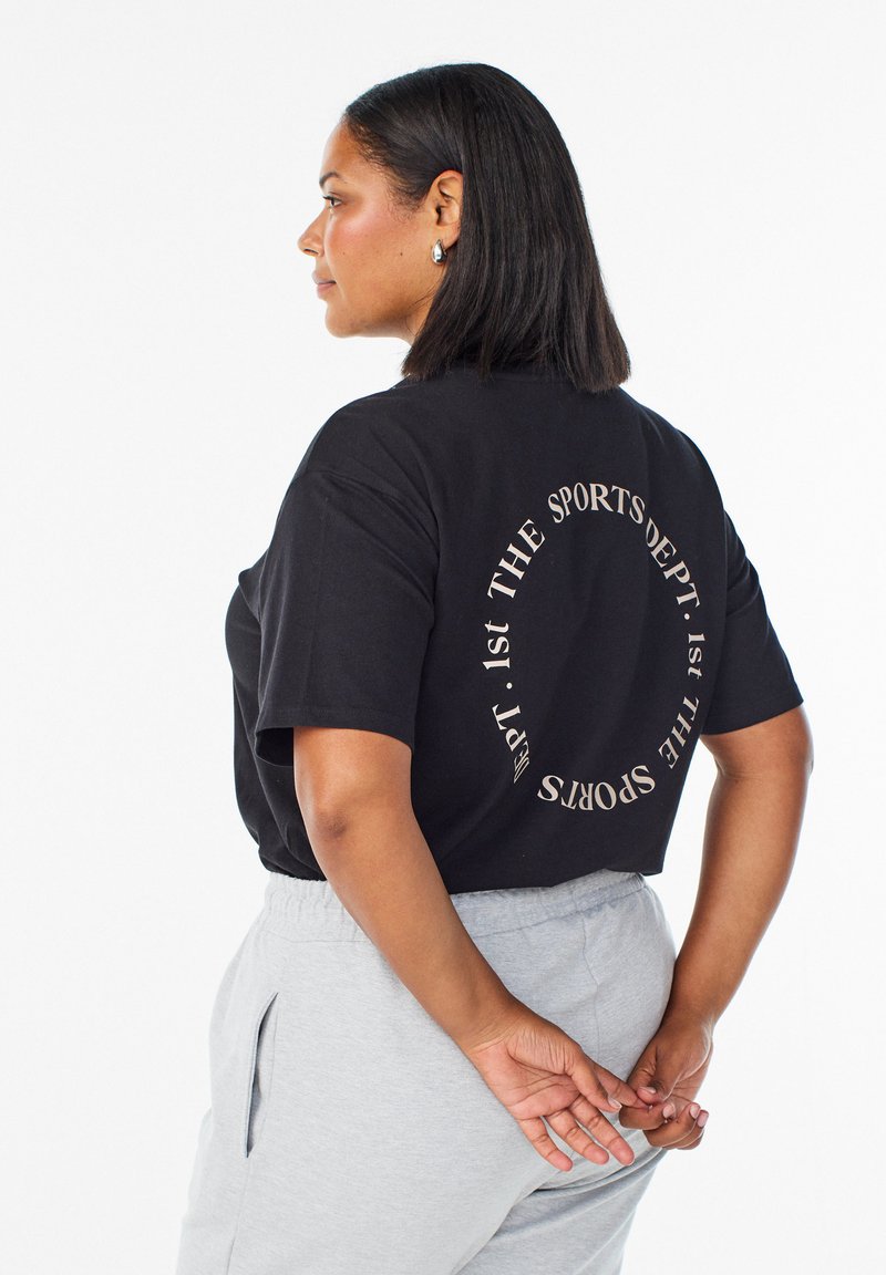 Sorth t-shirt med et rundt logo i beige tekst på ryggen. Materialet er bomuld med en afslappet pasform. Kombinationen af t-shirten med lysegrå sweatpants.