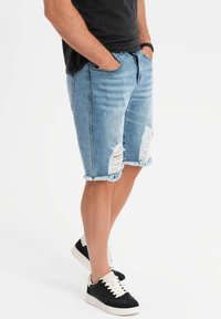 Pantalones cortos de mezclilla en azul claro con detalles desgastados y dobladillos deshilachados, combinados con zapatillas bajas en blanco y negro. Ajuste casual.