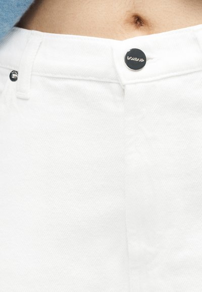 Κοντινή λήψη της μέσης και του κουμπιού λευκού τζιν παντελονιού με το όνομα της μάρκας, που δείχνει μέρος του γυμνού μεσαίου τμήματος και τον αφαλό.