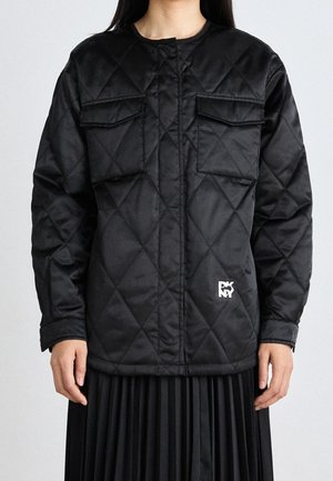 Personne portant une veste matelassée noire avec des poches à rabat sur la poitrine et un petit logo blanc près de l'ourlet inférieur, associée à une jupe noire plissée.