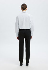 Camisa blanca de manga larga con pliegue en la parte trasera, combinada con pantalones negros a medida. El conjunto presenta un corte ajustado y una textura de tejido suave.