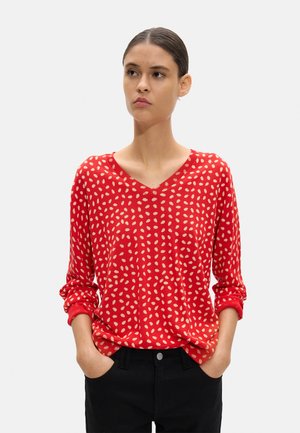 Kobieta ubrana w czerwony sweter z dekoltem w serek z beżowymi owalnymi wzorami oraz czarne spodnie, stojąca z rękami w kieszeniach, lekko patrząca w bok.