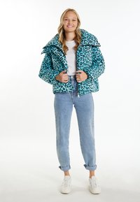 Tyrkysová péřová bunda s černým leopardím potiskem, přeplněný límec a zapínání na zip, doplněná světlými džíny s ohrnutými nohavicemi a teniskami.