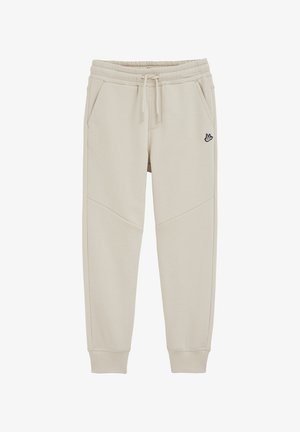 Pantalon de survêtement beige clair en tissu doux, avec une taille élastique, des poches latérales et un petit logo sur la cuisse gauche.