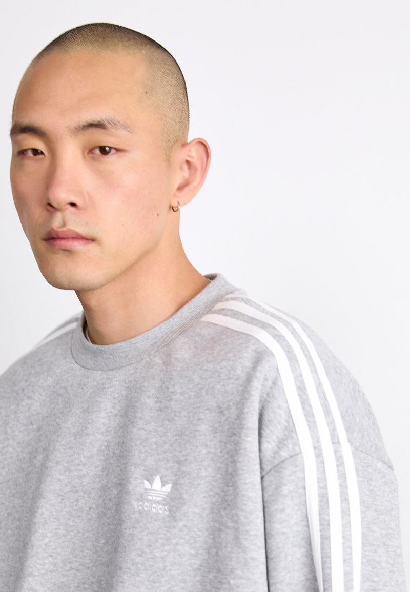 Sweatshirt gris en mélange de coton avec un design à trois bandes blanches sur les épaules. Présente un col rond et un petit logo sur la poitrine.