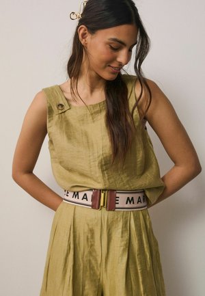 Woman wearing sleeveless olive-green linen dress with pleated skirt, a wide belt with pink and brown details, and a gold hair clip.