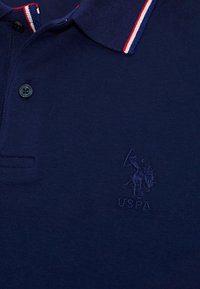 Námornícky modré polo tričko s golierom so pruhmi v červenej, bielej a modrej farbe, čiernymi gombíkmi a vyšitým logom hráča polo USPA na hrudi.