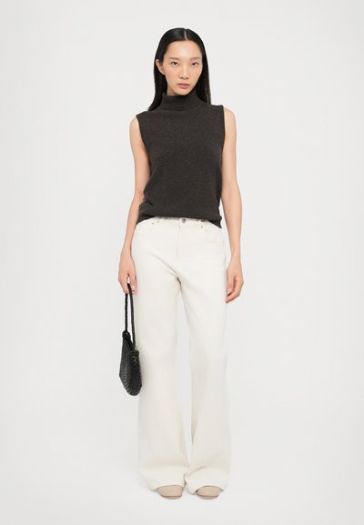 Tamna bezrukava turtleneck pulover uparen s širokim bijelim hlačama. Model drži malu crnu pletenu torbicu. Mekane teksture i neutralne boje.
