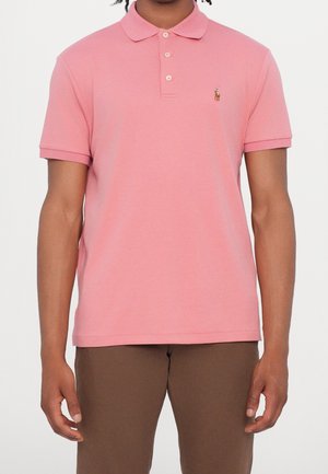Homme portant un polo rose à manches courtes avec un petit logo brodé sur la poitrine et un pantalon marron.