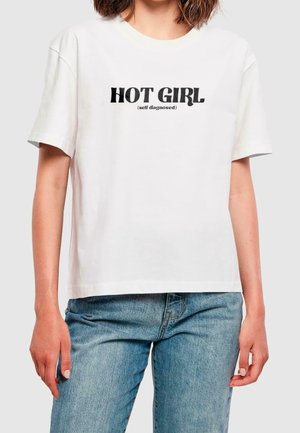 Kobieta w białej koszulce z czarnym napisem „HOT GIRL (samozdiagnozowana)”, w połączeniu z jasnoniebieskimi jeansami, na jednolitym tle.