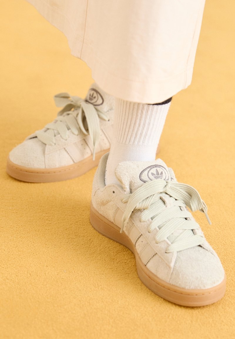 Baskets blanches Adidas avec semelles beige clair portées avec des chaussettes blanches côtelées et un pantalon couleur crème sur un sol jaune texturé.