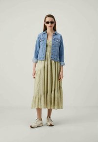 Kvinne i lys grønn midikjole, lyseblå denimjakke med oppbrettede ermer, hvite joggesko og mørke solbriller, som står mot en ensfarget bakgrunn.