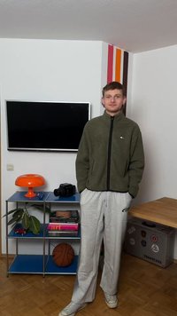 Junger Mann in grüner Jacke und grauer Jogginghose steht in einem Raum mit Fernseher, orangefarbener Lampe, Basketball, Kamera und Büchern im blauen Regal.