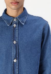 Camicia di jeans blu, con colletto a punta, chiusure con bottoni di metallo argentato e vestibilità classica. La superficie presenta una leggera texture e un colore uniforme.