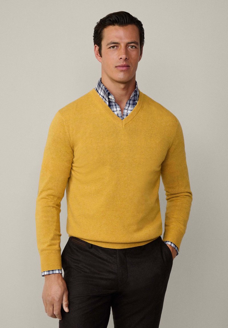 Pull jaune en v en maille, porté par-dessus une chemise à carreaux bleue et blanche. Pantalons à motifs sombres. Design simple et ajusté.