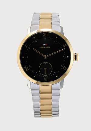 Montre-bracelet Tommy Hilfiger avec cadran noir, lunette et maillons centraux dorés, et bracelet métallique argenté et doré.