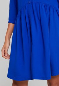 Robe bleue à manches courtes avec un corsage boutonné et une jupe froncée, fabriquée en tissu lisse avec un design fluide et un ourlet.
