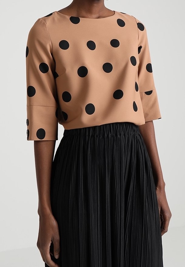 Brun blus med stora svarta prickar, trekvartslånga ärmar med knapppdetaljer, i kombination med en hög midja, plisserad svart kjol.