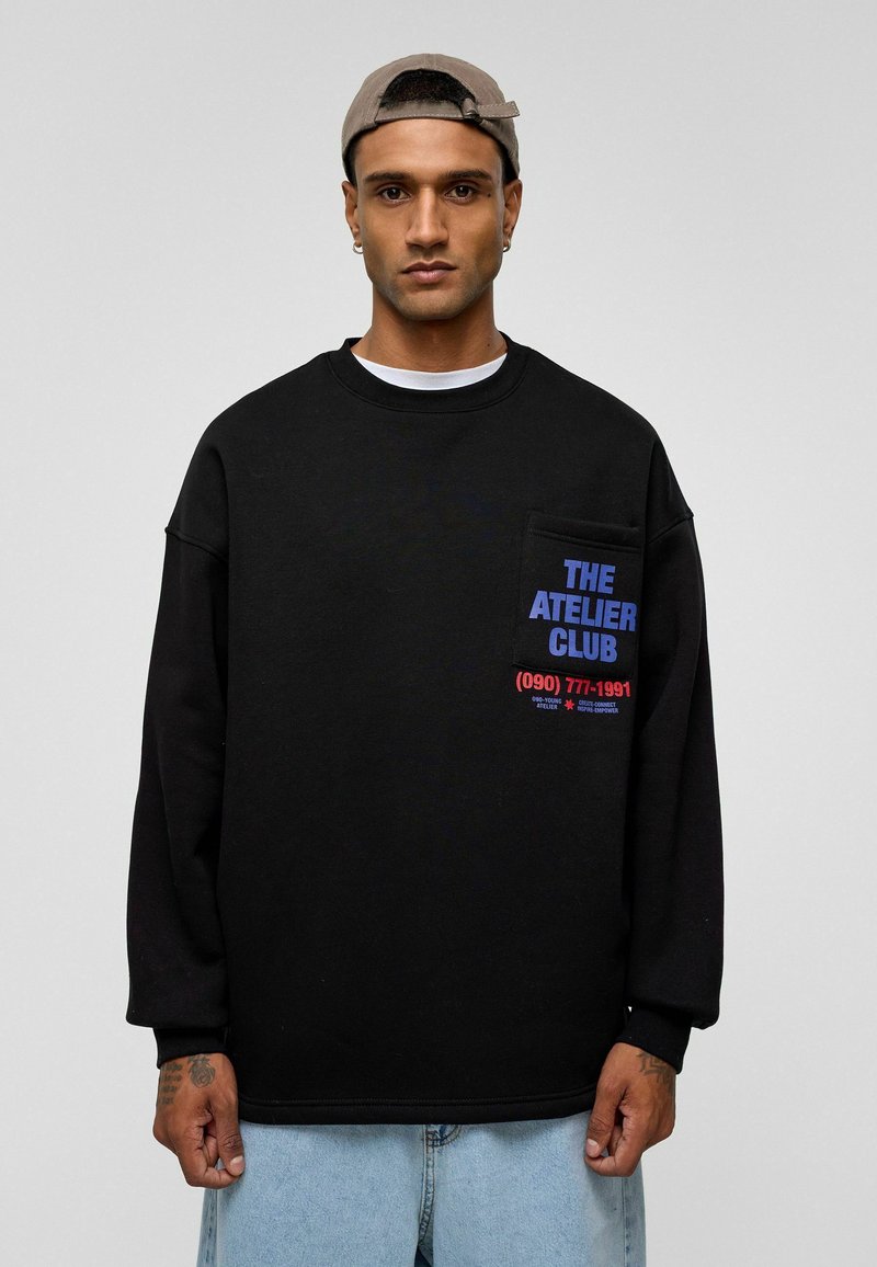 Zwarte sweatshirt met een ronde hals, lange mouwen en een voorkraag. Bevat blauwe en rode tekst, gemaakt van zachte stof. G gedragen met lichtblauwe jeans.