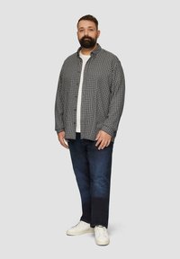 Geruit zwart-wit overhemd over een wit T-shirt, gecombineerd met donkerblauwe spijkerbroek en witte sneakers. Staand voor een lichte achtergrond.