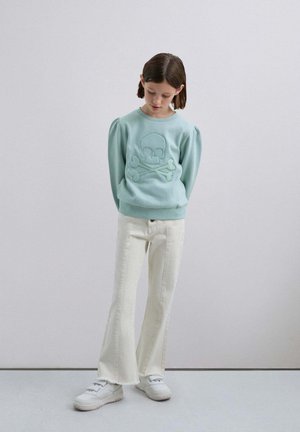 Sudadera azul claro con mangas abullonadas que presenta un bordado de calavera y huesos cruzados. Combinada con jeans blancos acampanados y zapatillas blancas.