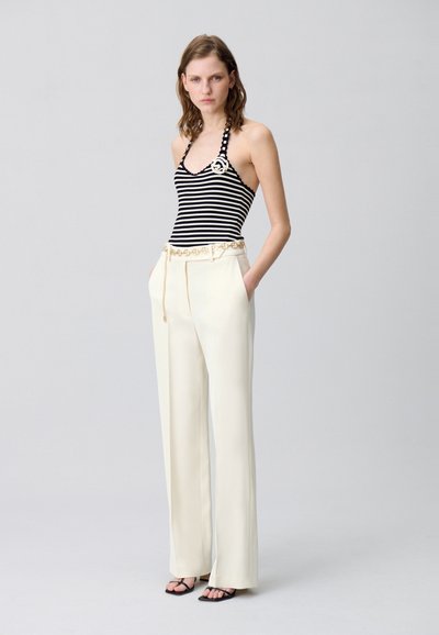 Top halter a strisce bianche e nere abbinato a pantaloni a gamba larga color avorio con cintura decorativa e tasche; la modella sta in piedi con le mani nelle tasche.