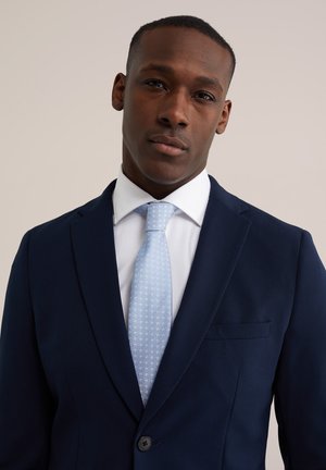 Man met een marineblauw colbert, wit overhemd en lichtblauwe stropdas met patroon, kijkt recht vooruit tegen een eenvoudige beige achtergrond.