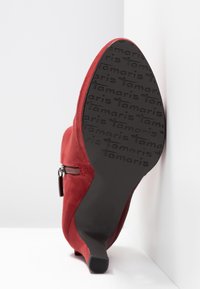 Bottine rouge en daim à talon avec un détail zipper, semelle estampillée du logo "Tamaris", photographiée par le dessous sur fond blanc.