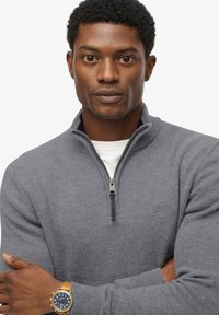 Maglione grigio con zip e colletto a coste, con logo sul petto. Il modello lo indossa sopra una camicia bianca, abbinato a un orologio.