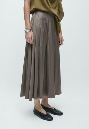 Žena v taupe plisované midi sukni, olivovém zavinovacím topu, stříbrném náramku a černých plochých botách stojící před jednobarevným pozadím.