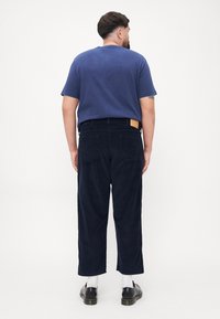 Navy corduroy pants with a straight-leg cut, back pockets, and a tan label on the waistband, paired with a blue t-shirt and black shoes.