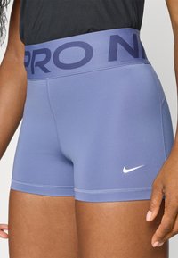 Skrócone spodenki Nike Pro w jasnoniebieskim kolorze z szerokim granatowym pasem z nazwą marki. Wykonane z gładkiego, elastycznego materiału z minimalną liczbą szwów.