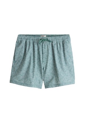 Hombre sin camiseta, con gafas de sol y bañador, está descalzo sobre una roca junto a la playa, sosteniendo una toalla beige alrededor de su cuello.
